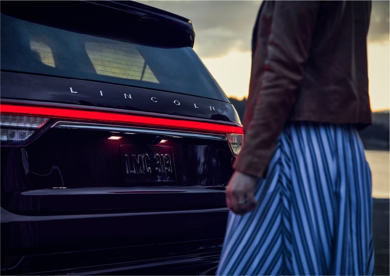 A person is shown near the rear of a 2023 Lincoln Aviator® SUV as the Lincoln Embrace illuminates the rear lights | Preston Lincoln in Hurlock MD