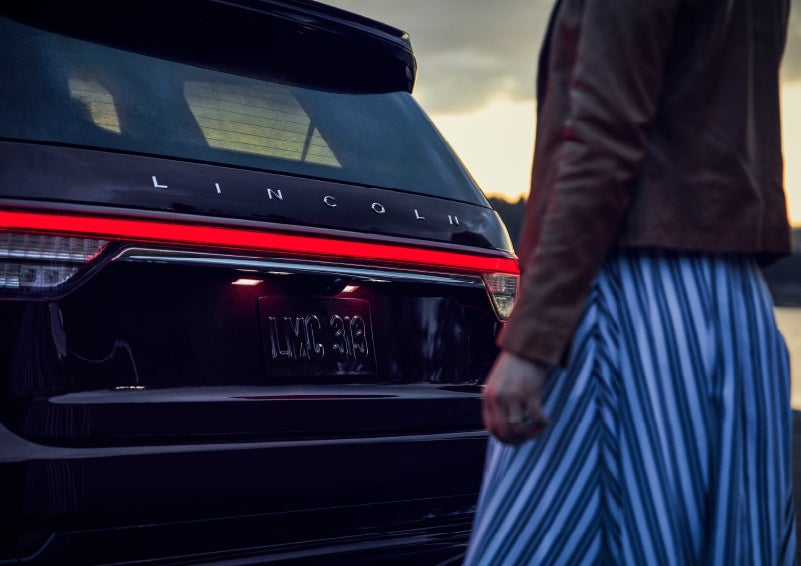 A person is shown near the rear of a 2024 Lincoln Aviator® SUV as the Lincoln Embrace illuminates the rear lights | Preston Lincoln in Hurlock MD